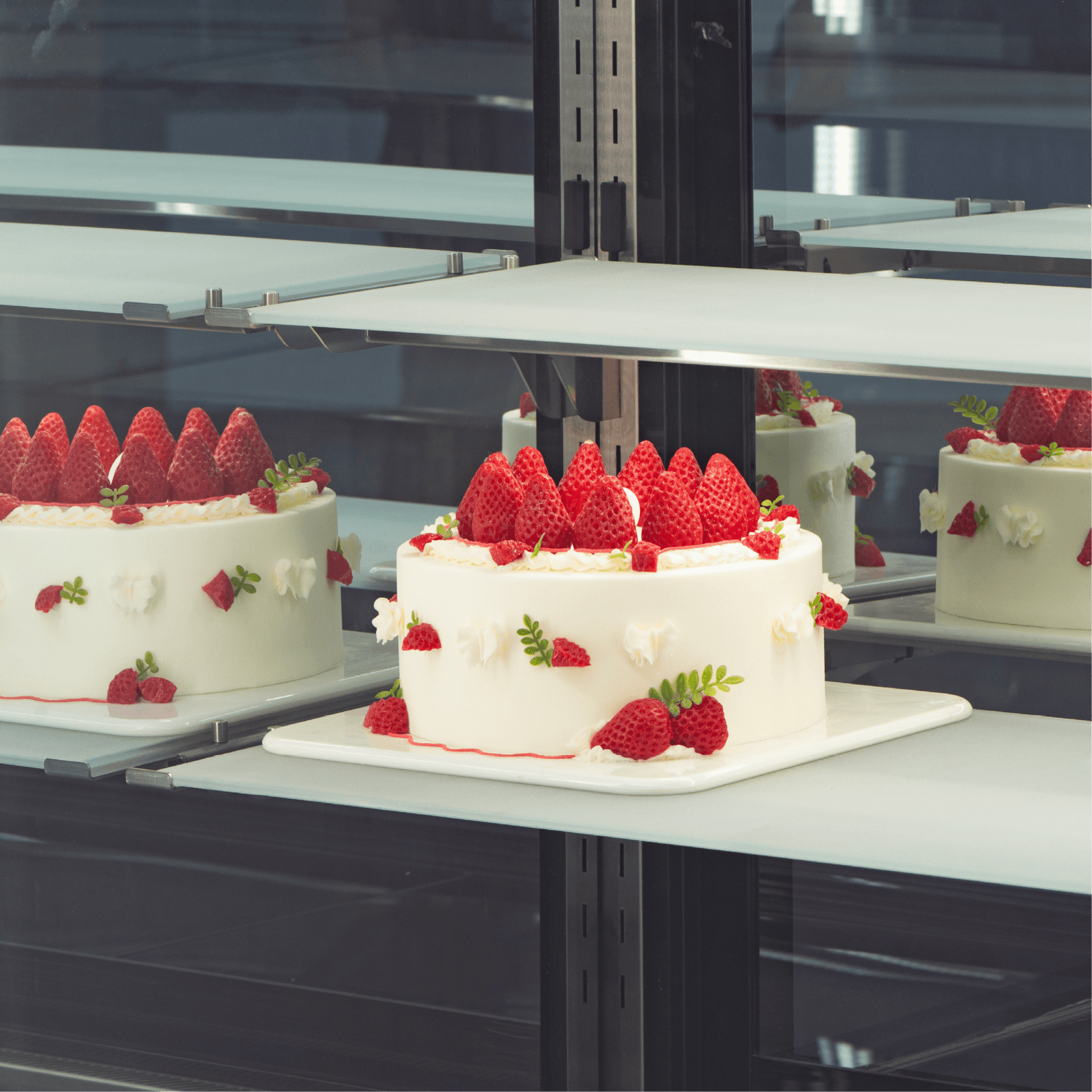 Four-tier refrigerated cake display with black stainless steel frame and clear glass shelves showcasing a white cake topped.