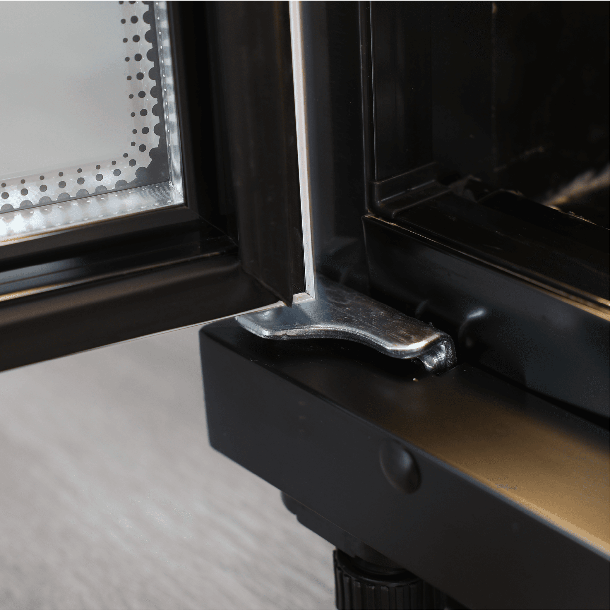 Close-up of black painted steel frame with glass door hinge on upright display refrigerator.