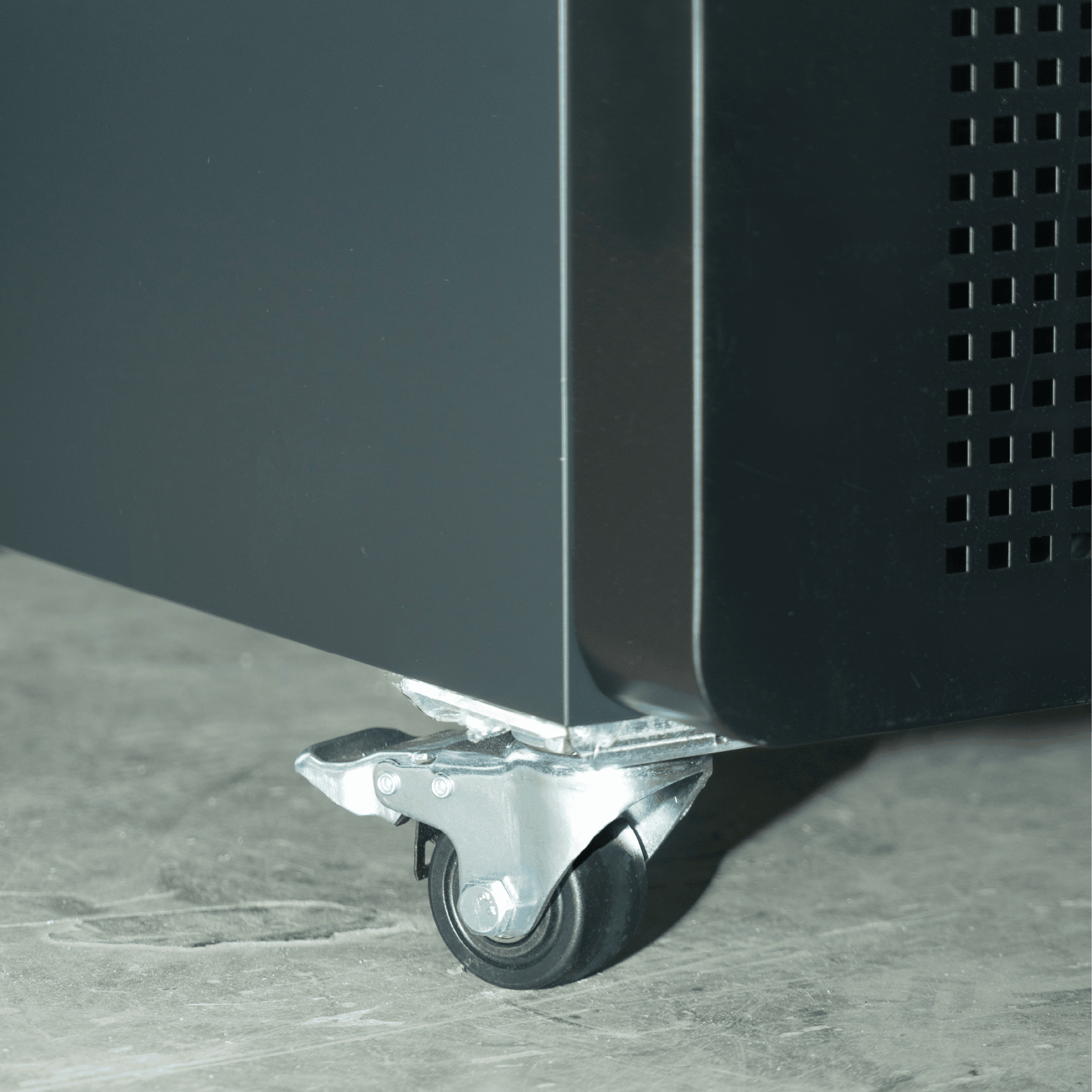 Close-up of black steel upright fridge corner with small silver caster wheel on gray floor.