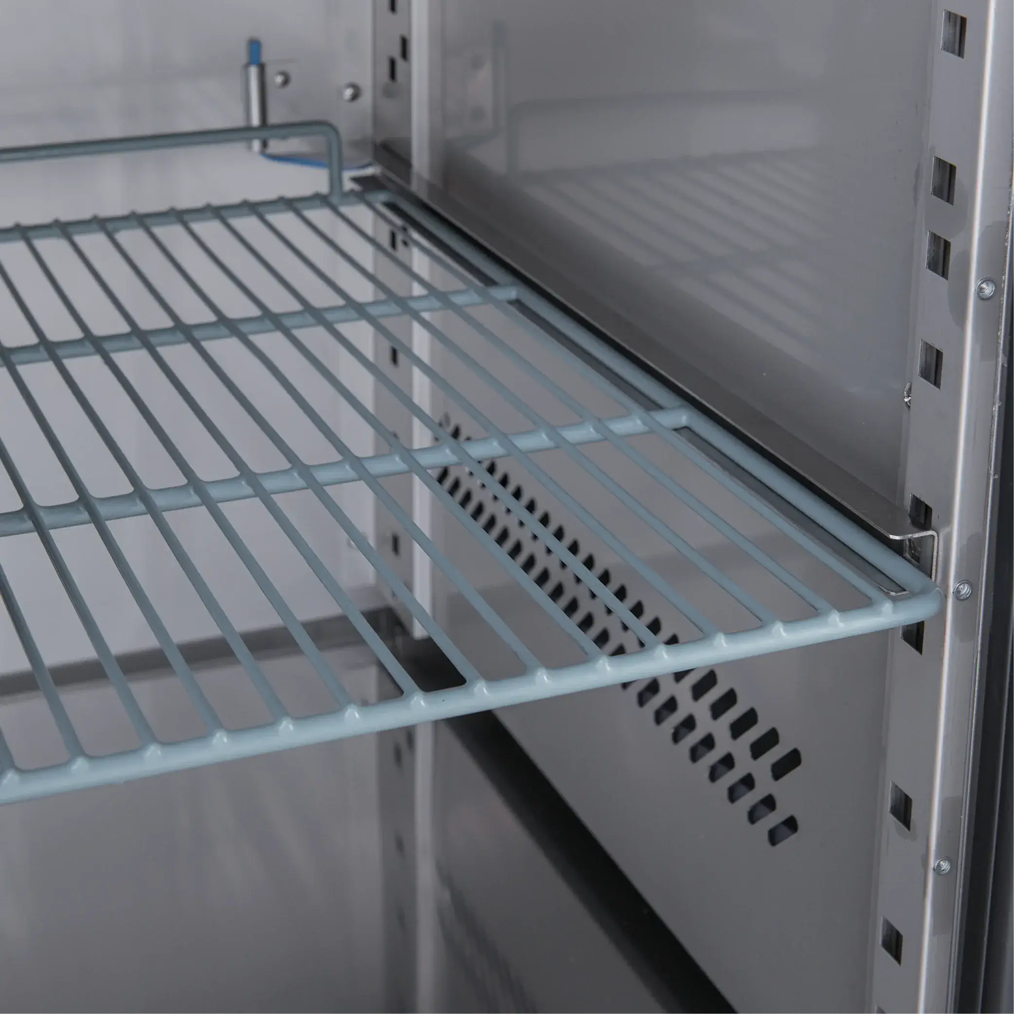 Stainless steel underbench fridge interior with adjustable wire shelves and ventilation panel.
