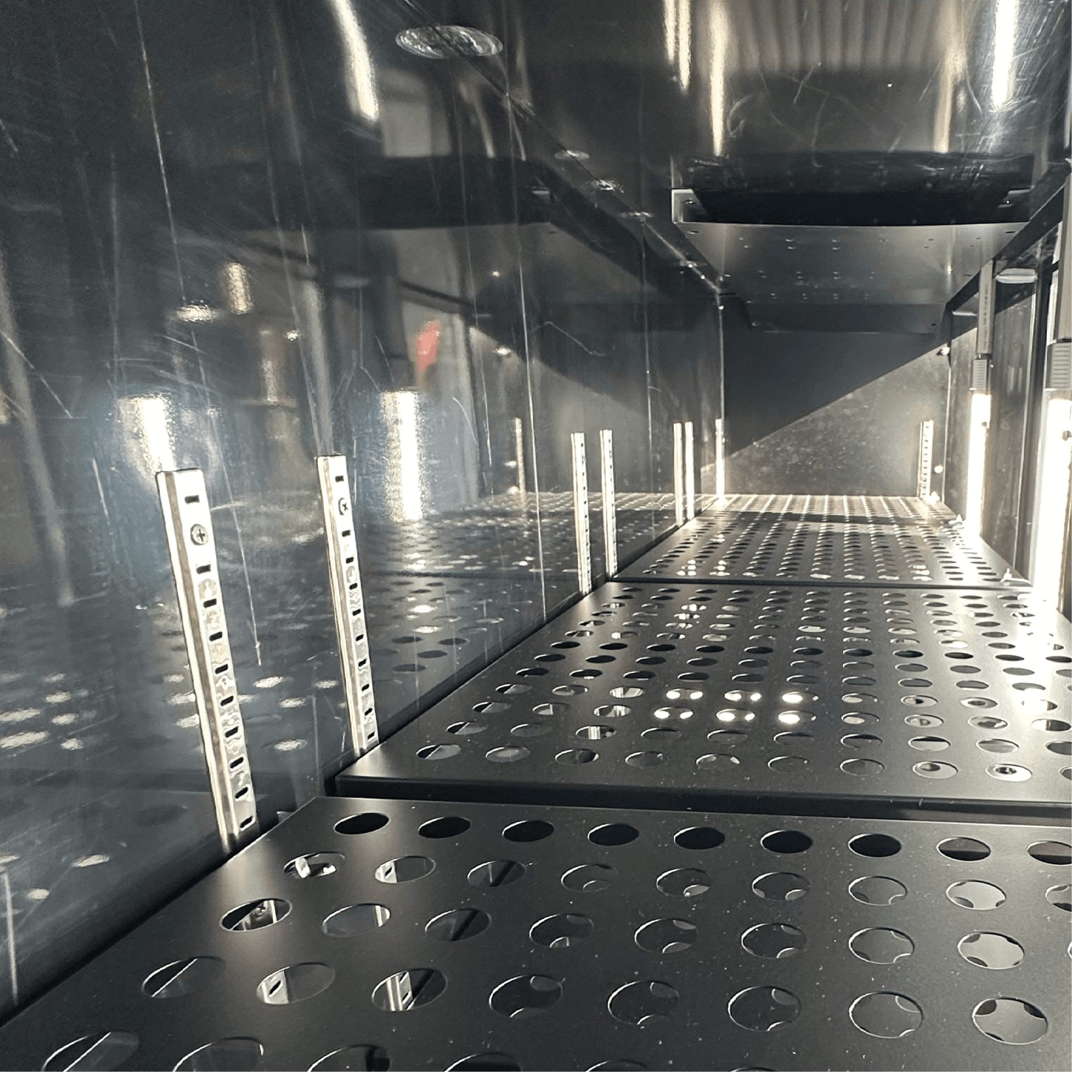 Black perforated metal shelves inside a stainless steel underbench refrigerator with adjustable shelf slots.