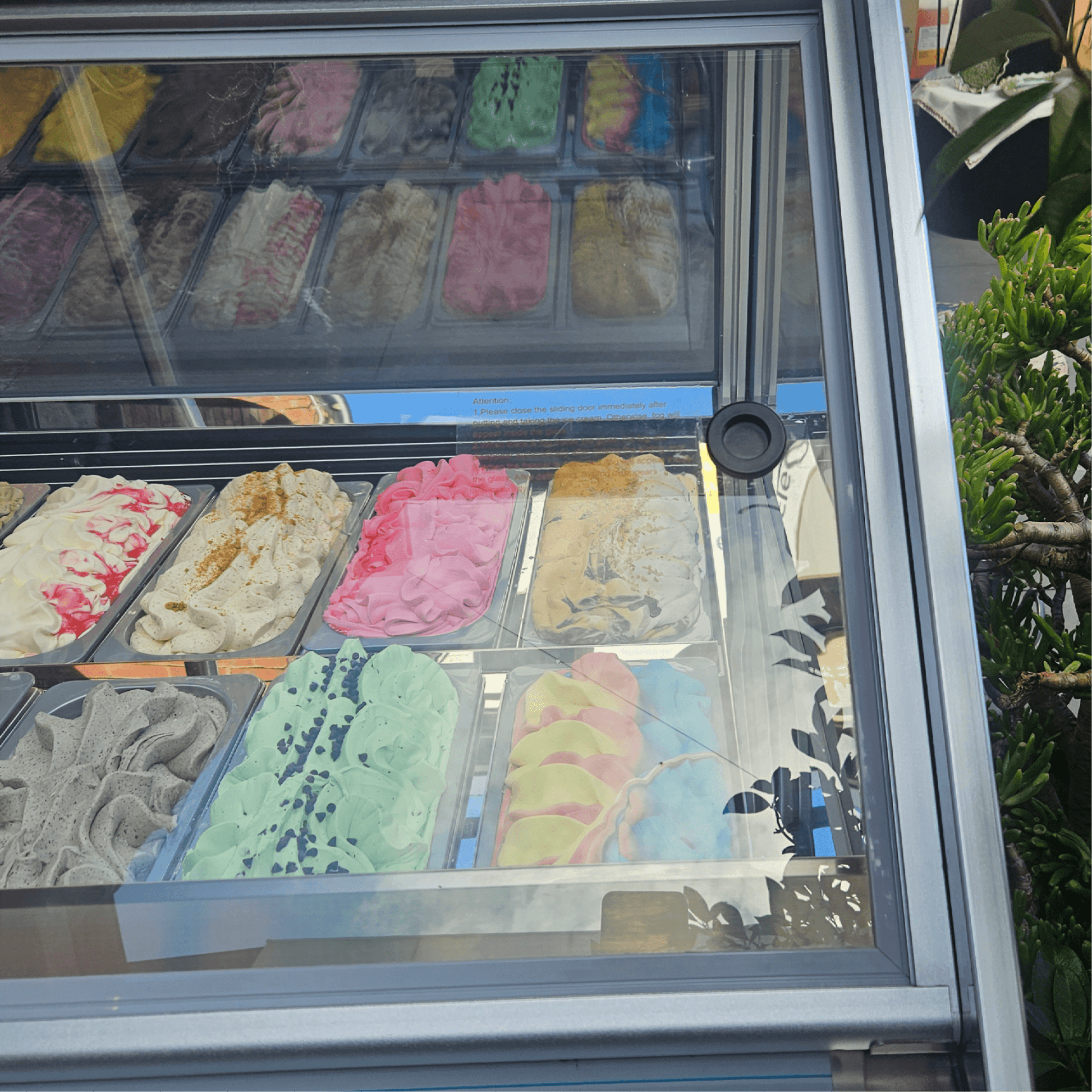 White painted steel and glass freezer with 16 rectangular tubs of assorted colorful ice cream flavors visible inside.