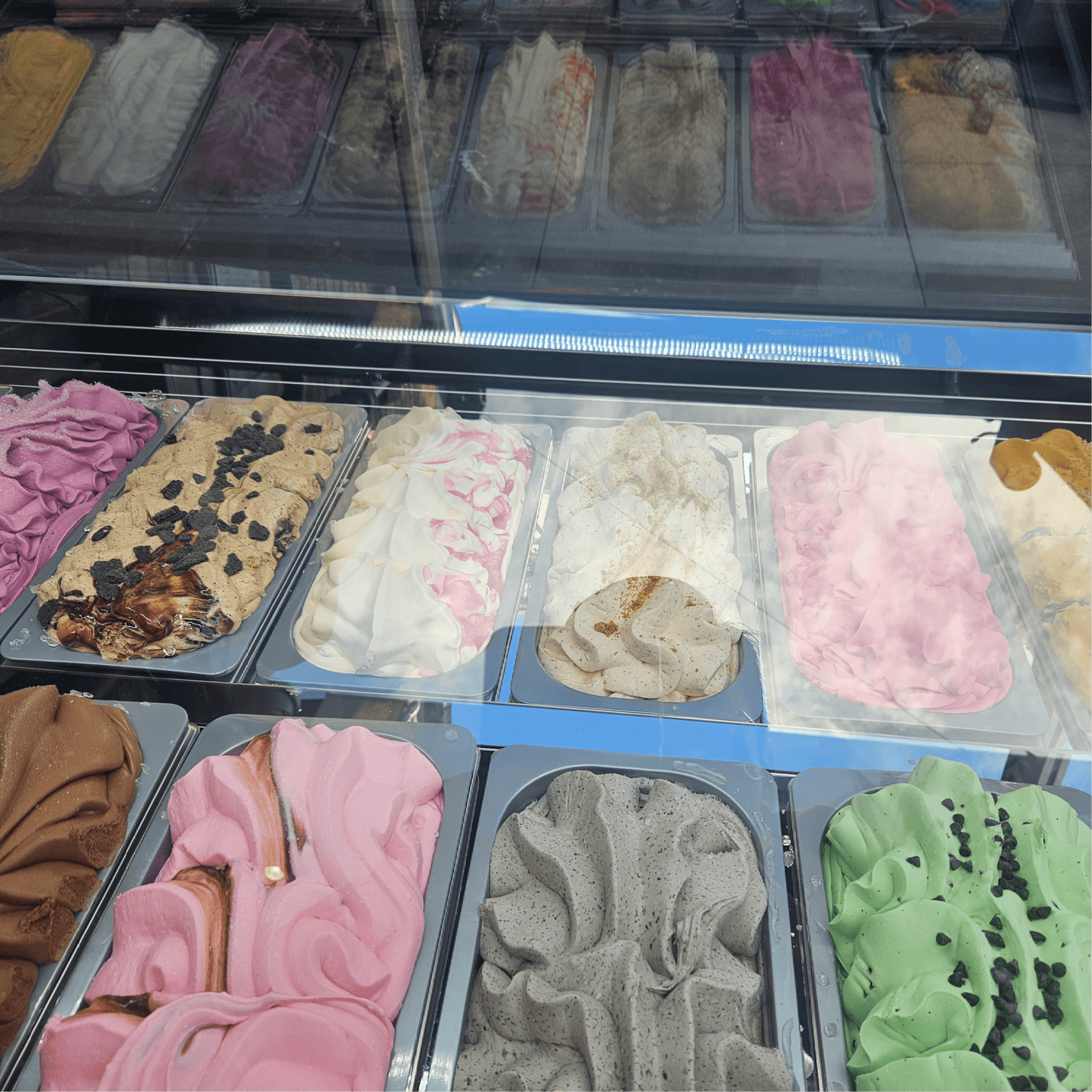 White steel and glass ice cream freezer with 18 rectangular tubs of assorted ice cream and gelato flavors including pink,.